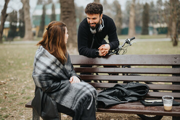 A man with headphones and a woman in a plaid coat enjoy a casual chat on a wooden park bench, with...