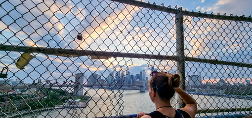 A woman with sunglasses on her head looking through a hole in a fence above Hudson River on a...