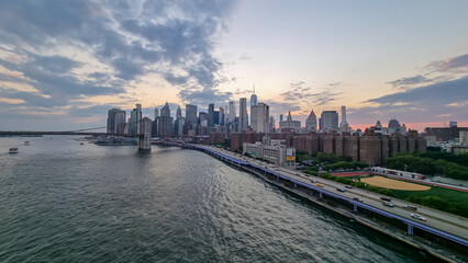 Fototapeta premium A view at the panorama of New York City's skyscrapers. Brooklyn Bridge in front is lit up. Overcast. Missing freedom. The sun is slowly setting behind the city. Busy and dazzling city.