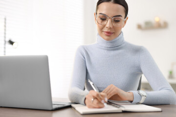 Young woman in glasses writing down notes during webinar at table in office