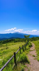 Obraz premium Vast mountain views stretch out from a dirt trail with a rustic wooden fence, under a brilliant blue sky and fluffy clouds