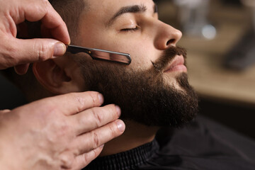 Professional barber shaving client's beard with blade in barbershop, closeup