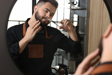 Handsome young man trimming mustache with scissors near mirror indoors