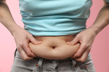 Woman touching belly fat on pink background, closeup. Overweight problem