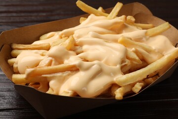 Tasty potato fries and cheese sauce in paper container on wooden table, closeup