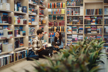 A young man and woman sit on the floor of a well-stocked library, engaged in a discussion over a...
