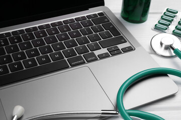 Laptop, stethoscope and pills on white wooden table, closeup