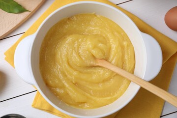 Delicious lemon curd in bowl and spoon on white wooden table, top view