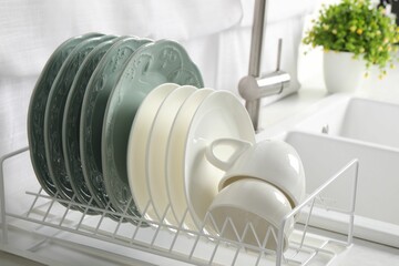 Drainer with different clean dishware and cups on white table in kitchen