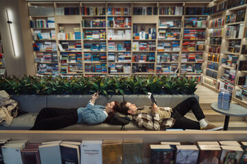 A young couple enjoys a quiet moment, lying down and reading books in a well-stocked modern library...