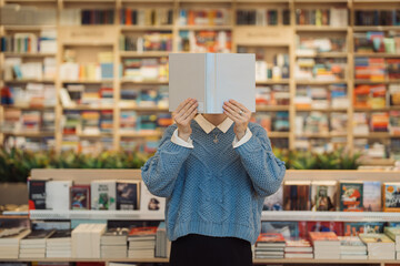 A young woman holds a book in front of her face cover, standing amidst a vibrant and varied...