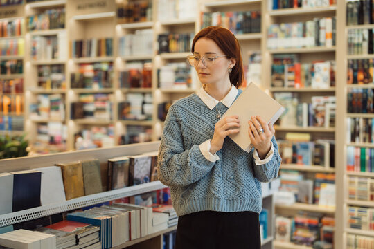 A thoughtful young woman in a cozy sweater holds a book in a well-stocked library, surrounded by shelves full of literature.