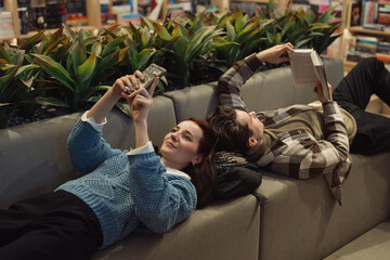 A young woman takes a selfie while a man reads a book beside her on a comfortable sofa, surrounded...