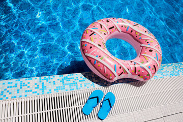 Pink inflatable ring and blue rubber flip-flops by blue outdoor pool water. Poolside relaxation.