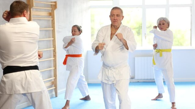 Diligent old man attendee of karate classes practicing kata standing in row with others in sports hall