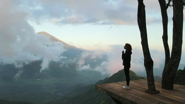 Silhouette of a young girl standing on a wooden platform high in the mountains, folding her hands in namaste and meditating, with a beautiful view of the clouds and the volcano mountain. Bali.
