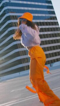 Vertical video. Joyful multi racial girl in sports outfit wearing trendy bright orange cargo pants and bucket hat dancing into music at city downtown. Hip-hop queen making twerk. Breakdancer woman