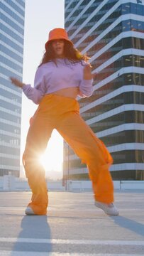Vertical video. Hip-hop queen making twerk. Breakdancer woman enjoying sunset 4K. Joyful multi racial girl in sports outfit wearing trendy bright orange cargo pants and bucket hat dancing into music