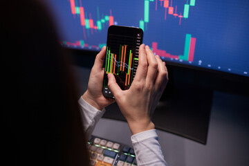 Oil prices moves up on mobile screen. Woman's hand holds a mobile phone. Monitor in the background.
