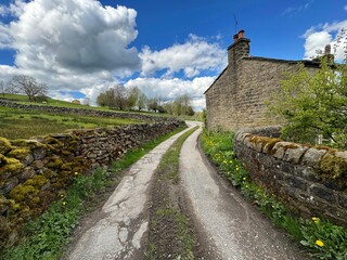 A country road winds past an old stone house, under a sky filled with white clouds, with dry stone walls and wild plants near, West Lane, Sutton-in-Craven, UK