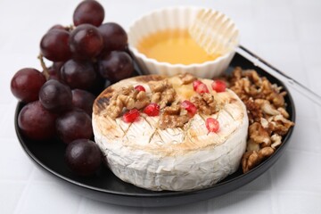 Plate with tasty baked camembert, honey, grapes, walnuts and pomegranate seeds on white tiled table, closeup