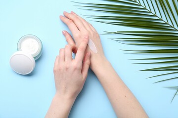 Woman applying hand cream on light blue background, top view