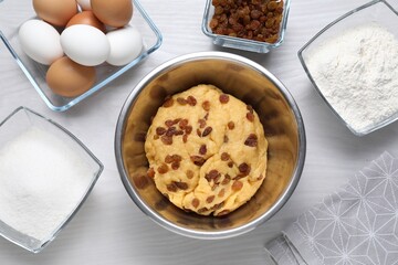 Dough with raisins in bowl and ingredients on white wooden table, flat lay