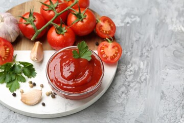Delicious ketchup in bowl, parsley, garlic and tomatoes on grey textured table, above view. Space for text