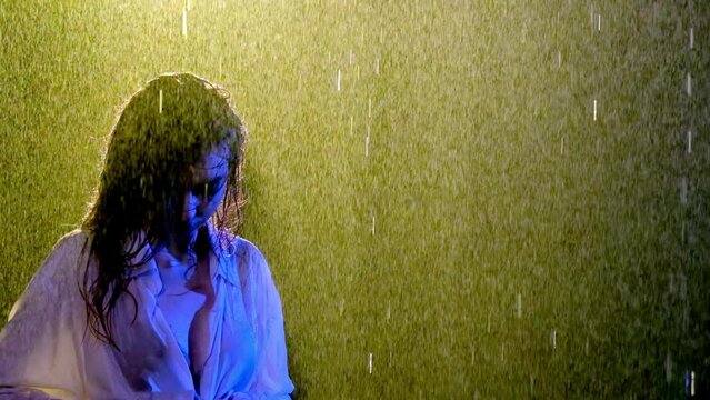 Lady in wet blouse stands with head bowed under streams of rain. Strong flow of water fully soaks woman with slender shoulders