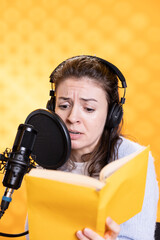 Woman with frown on face wearing headphones reading aloud from book into mic against background....