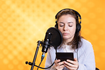 Narrator wearing headphones reading aloud from ebook on ereader into professional microphone...