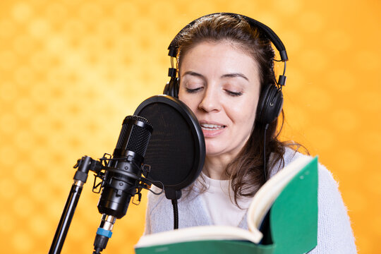 Smiling lady doing voiceover reading of book to produce audiobook. Happy voice actor using storytelling skills to entertain audience while producing digital recording of novel, studio background