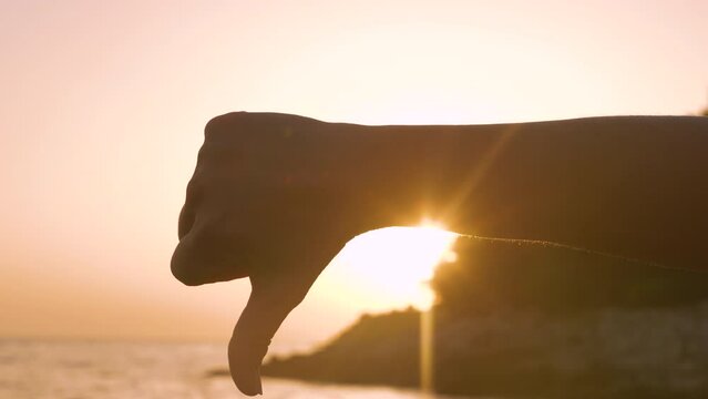 LENS FLARE, SILHOUETTE: Golden lit thumbs down gesture on the beach at sunset. Close up view of a familiar hand gesture symbolizing disapproving of the premature end of summer vacations by the sea.