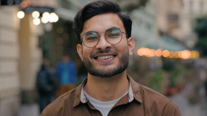 Close up portrait happy wide toothy smiling successful young handsome Indian Arabian Latino ethnic male student man guy businessman glasses posing looking friendly at camera outdoors city urban street