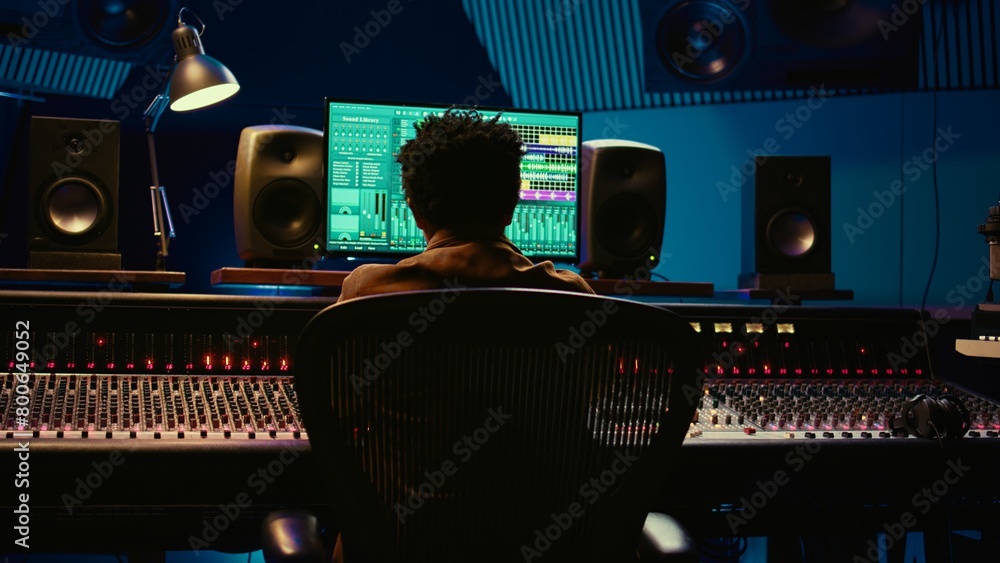 Poster African american sound designer mixing and mastering tracks in professional studio, using audio console with moving faders and colored meters. Music producer working on control room. Camera A.