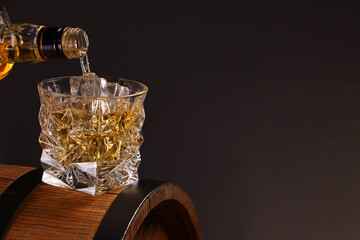 Pouring whiskey from bottle into glass on wooden barrel against dark background, closeup. Space for...