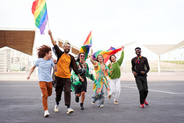 Wide shot of a group of people celebrating the pride day, holding rainbow flags while running excited and looking at camera cheerfully. Copy space.