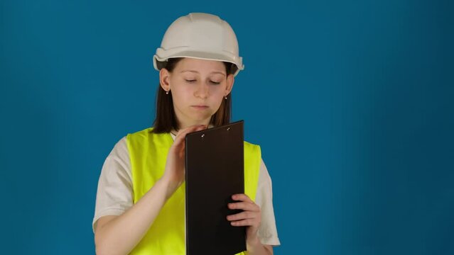 Serious young woman taskmaster with clipboard observes workplace on blue background. Safety inspector controls construction site work
