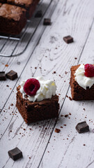 Chocolate chip brownies with whipped cream and raspberries on a wooden background with crumbs and dark chocolate pieces around. Selective focus.