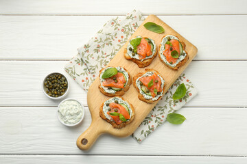 Tasty canapes with salmon, capers, cucumber and sauce on white wooden table, top view