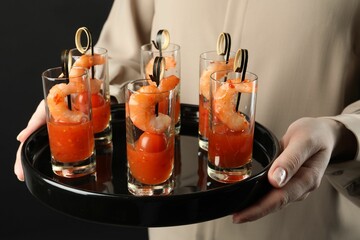 Woman holding tasty canapes with shrimps, tomatoes and sauce in shot glasses on black background,...