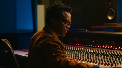 Portrait of african american audio technician uses mixing console with knobs and sliders in control...