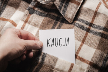  White card with a handwritten inscription "Kaucja", held in the hand against the background of a brown plaid shirt (selective focus), translation: deposit