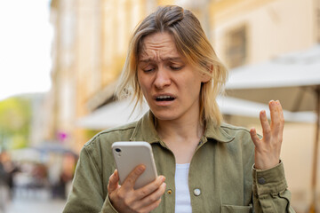 Caucasian young woman using smartphone typing browsing loses becoming surprised sudden lottery results bad news fortune loss app fail virus thief fraud outdoors. Tourist girl on urban city street.