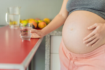 Embracing the vital benefits of water during pregnancy, a pregnant woman stands in the kitchen with a glass, highlighting hydration's crucial role in maternal well-being