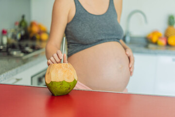 Quenching her pregnancy thirst with a refreshing choice, a pregnant woman joyfully drinks coconut water from a coconut in the kitchen, embracing natural hydration during this special journey
