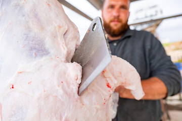 The butcher is cutting meat outdoors, holding his knife, and smiling with a bearded face