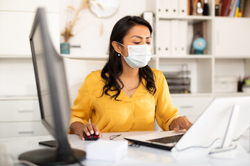 Portrait of confident Latina wearing medical mask working on computer in office. New life reality...