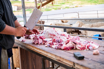 The butcher cuts the meat with knife on wooden cutting board.Close-up piece of raw meat with a chef's knife outdoors in springfield with sheeps barn  in the background - obrazy, fototapety, plakaty
