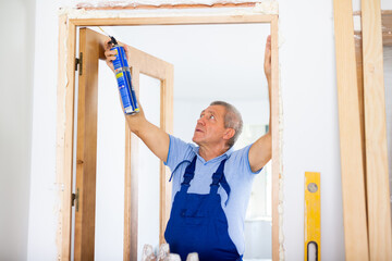 Focused middle-aged contractor repairman in a blue uniform filling door jambs with polyurethane...